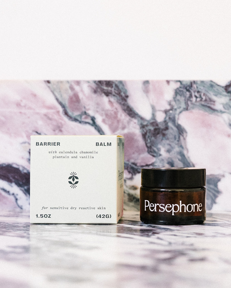 Barrier Balm by Persephone shown with its yellow product box and the dark amber jar with a black lid, set against a pink and gray marble background.