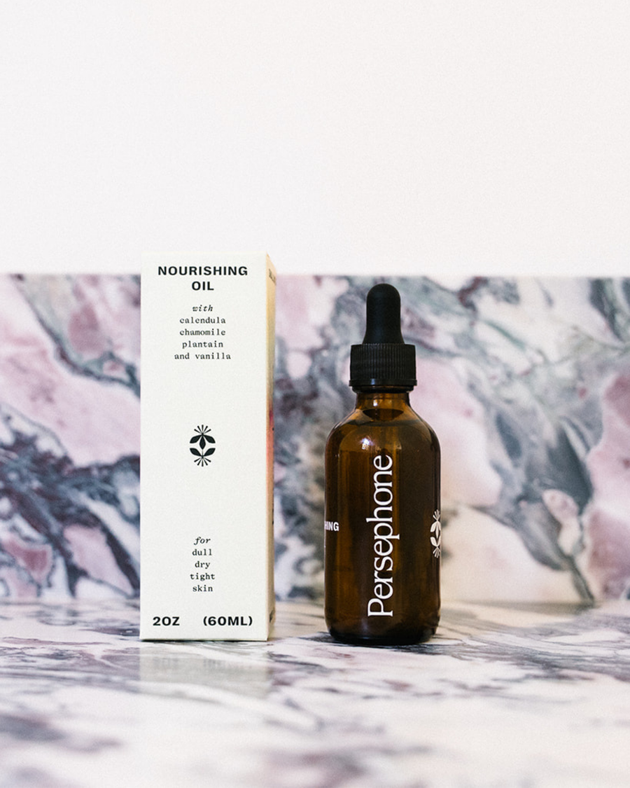 A 2 oz amber glass dropper bottle of Persephone Nourishing Oil next to its yellow cardboard packaging box, set against a marble-patterned background.
