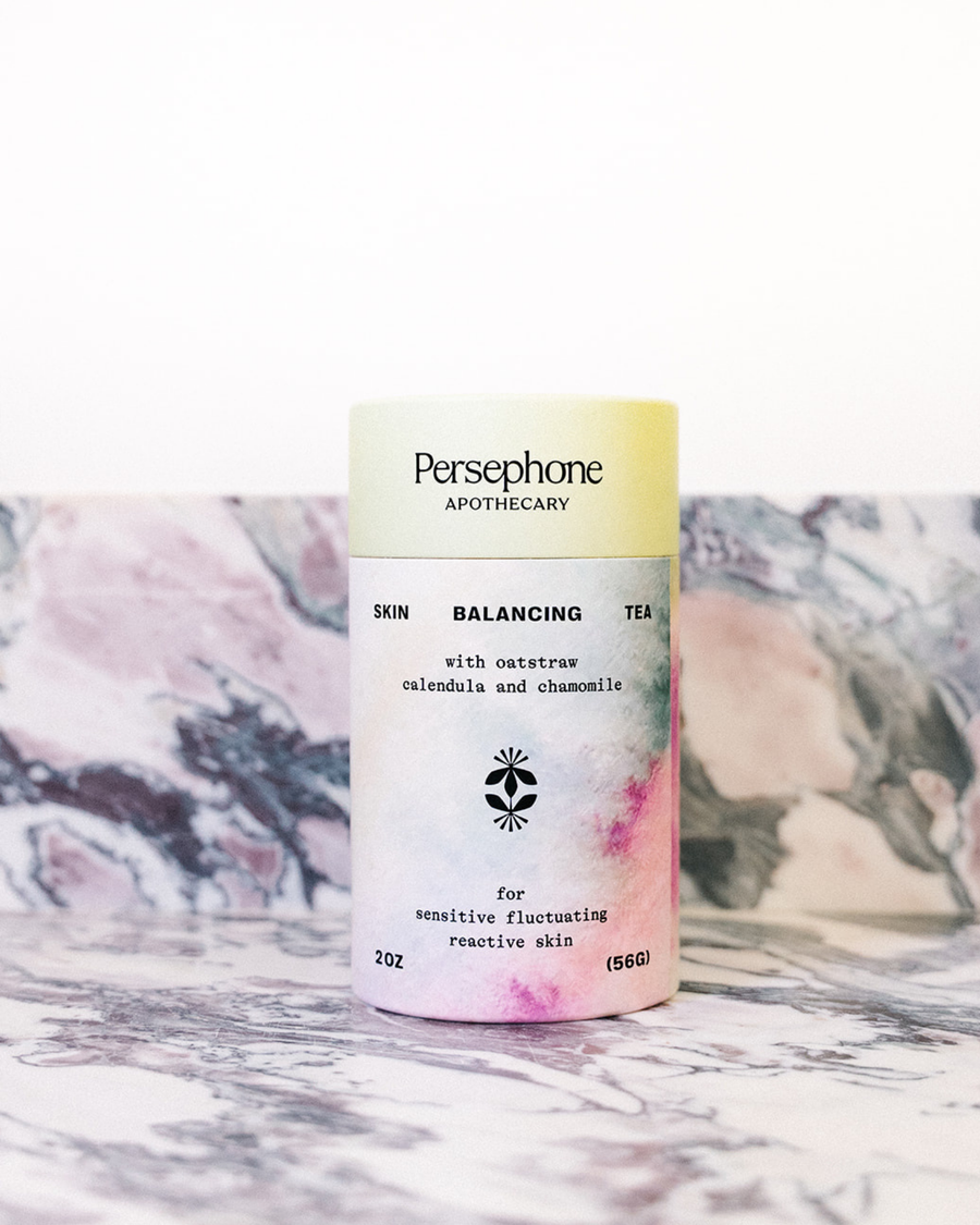 Skin Balancing Tea by Persephone shown in its pastel gradient canister next to its box, set against a pink and gray marble background.